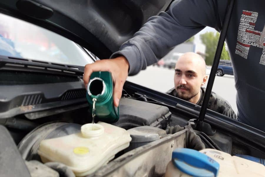 Man observing his friend as he pours oil on a car engine